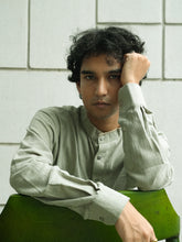 Load image into Gallery viewer, Man in a moss green shirt sitting on a green chair against a white tiled wall.