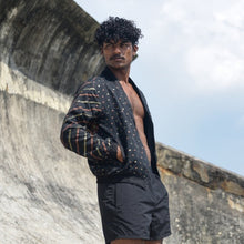 Load image into Gallery viewer, Man wearing a patterned cardigan over a black shirt and shorts standing against a textured wall with blue sky.