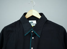 Load image into Gallery viewer, Black shirt with teal stitching on a wooden hanger against a grass background
