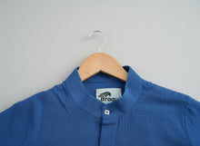 Load image into Gallery viewer, Blue shirt on a wooden hanger with a brand label against a grass background
