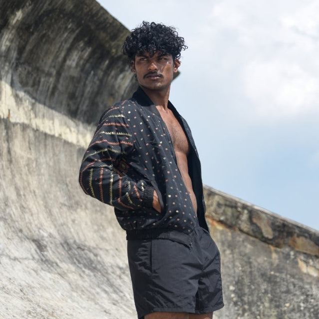 Man wearing a patterned cardigan over a black shirt and shorts standing against a textured wall with blue sky.